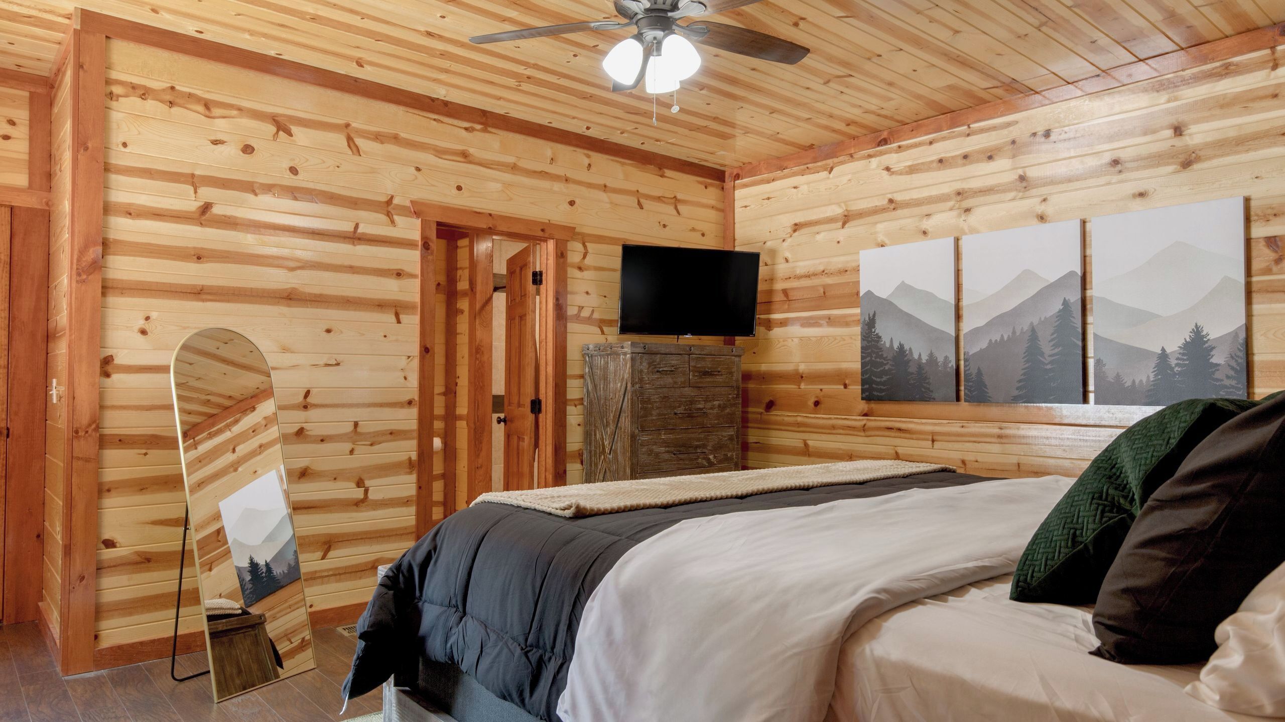 Un dormitorio de cabaña de madera con cama, ventilador de techo, televisor y una pintura con vistas a la montaña.