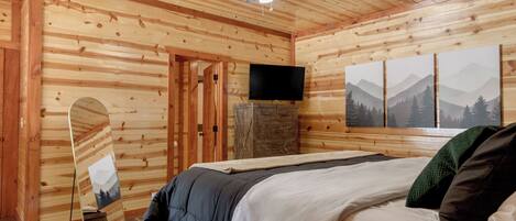 Un dormitorio de cabaña de madera con cama, ventilador de techo, televisor y una pintura con vistas a la montaña.