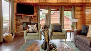 Un acogedor interior de cabaña con chimenea de piedra, dos sillones verdes, un sofá marrón y un televisor montado en la pared.