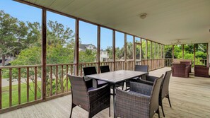 A covered outdoor patio with a table and chairs, large windows, and a view of greenery and houses. - Ion by Avantstay Walk to Beach, Dining, Shopping Central Location Deck (Sullivan's Island)