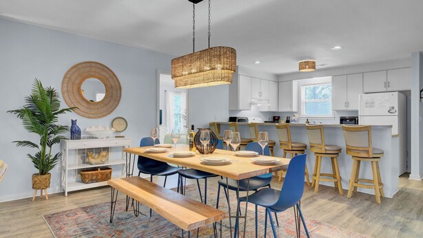 A modern kitchen with white cabinets, a dining area with a wooden table and blue chairs, and a hanging wicker light fixture. - Ion by Avantstay Walk to Beach, Dining, Shopping Central Location Deck (Sullivan's Island)