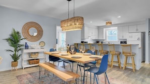 A modern kitchen with white cabinets, a dining area with a wooden table and blue chairs, and a hanging wicker light fixture. - Ion by Avantstay Walk to Beach, Dining, Shopping Central Location Deck (Sullivan's Island)