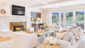 A modern living room with a fireplace, comfortable seating, a flat-screen TV, and a view through glass doors to an outdoor area. - La Jolla Living by Avantstay A+ Location Private Backyard Lounge (La Jolla)