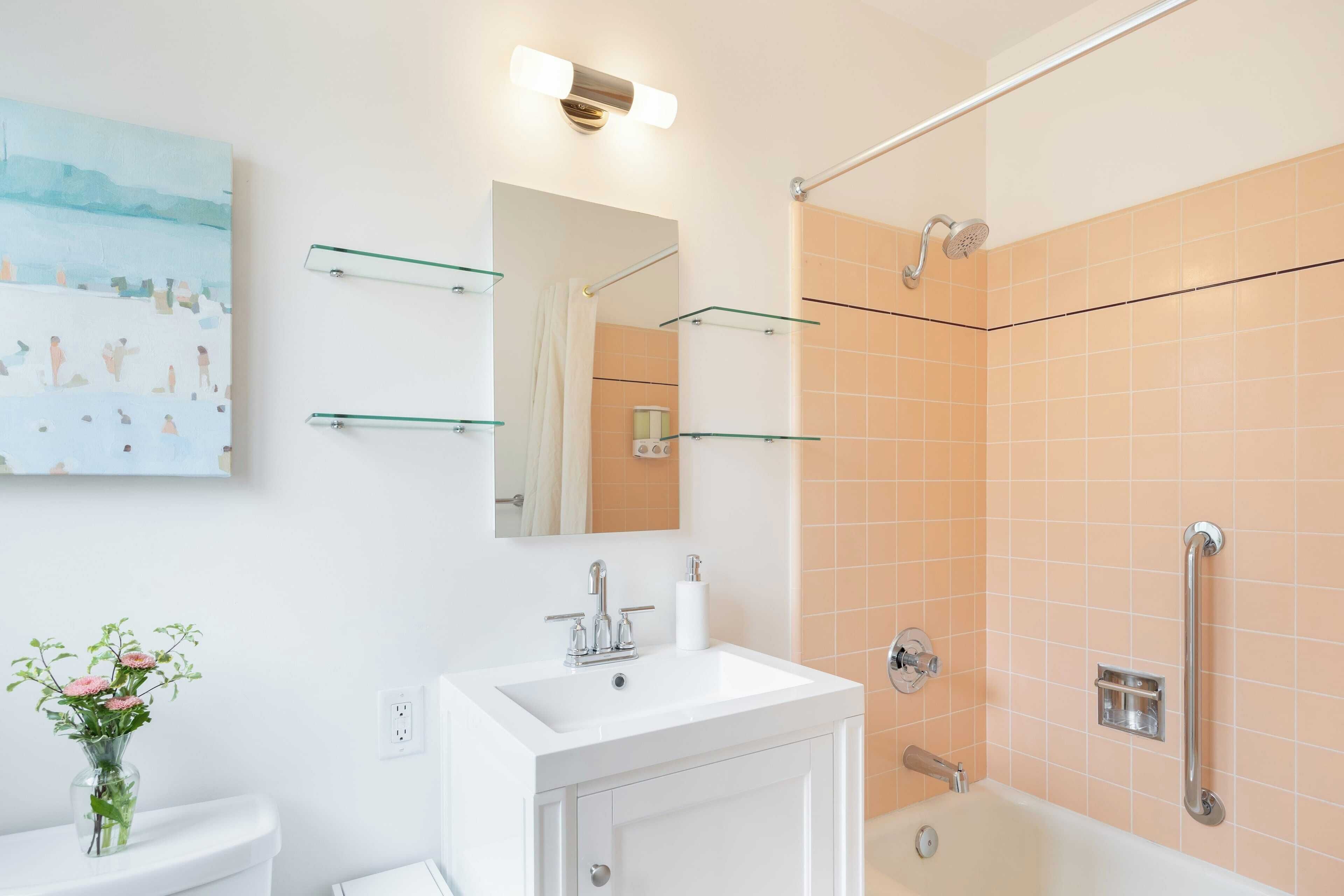 A bathroom with a white sink, mirror, and glass shelves; a shower with a handheld showerhead; and a toilet with a vase of flowers.