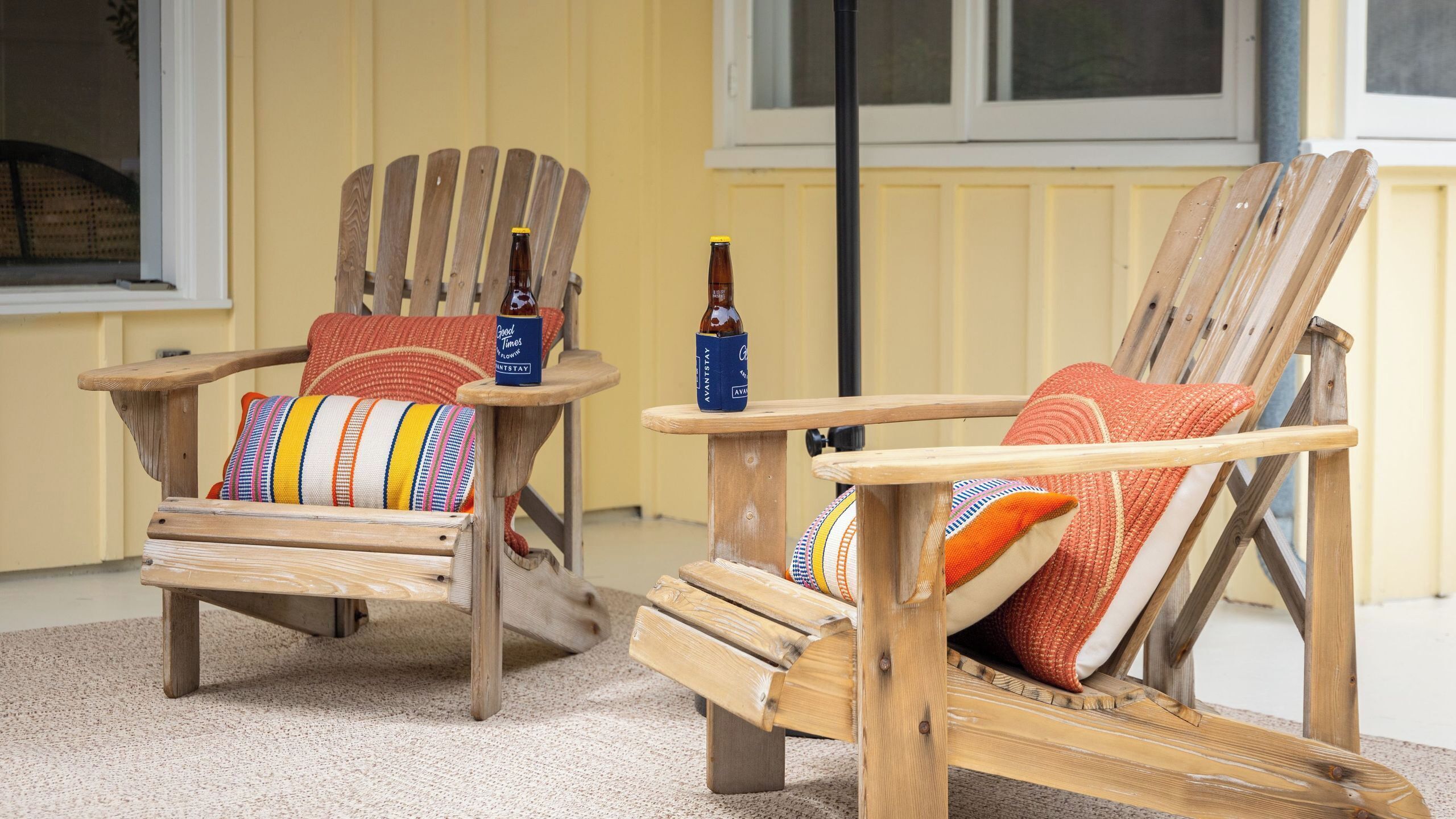 Two wooden Adirondack chairs with colorful cushions, a bottle of beer, and a bottle of wine on a small wooden table.