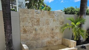 Une cabine de douche en pierre avec un banc, entourée d’un petit muret et de palmiers.