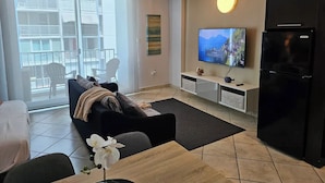 Un salon moderne comprenant un canapé, un téléviseur, un réfrigérateur et un balcon aménagé avec du mobilier d’extérieur.