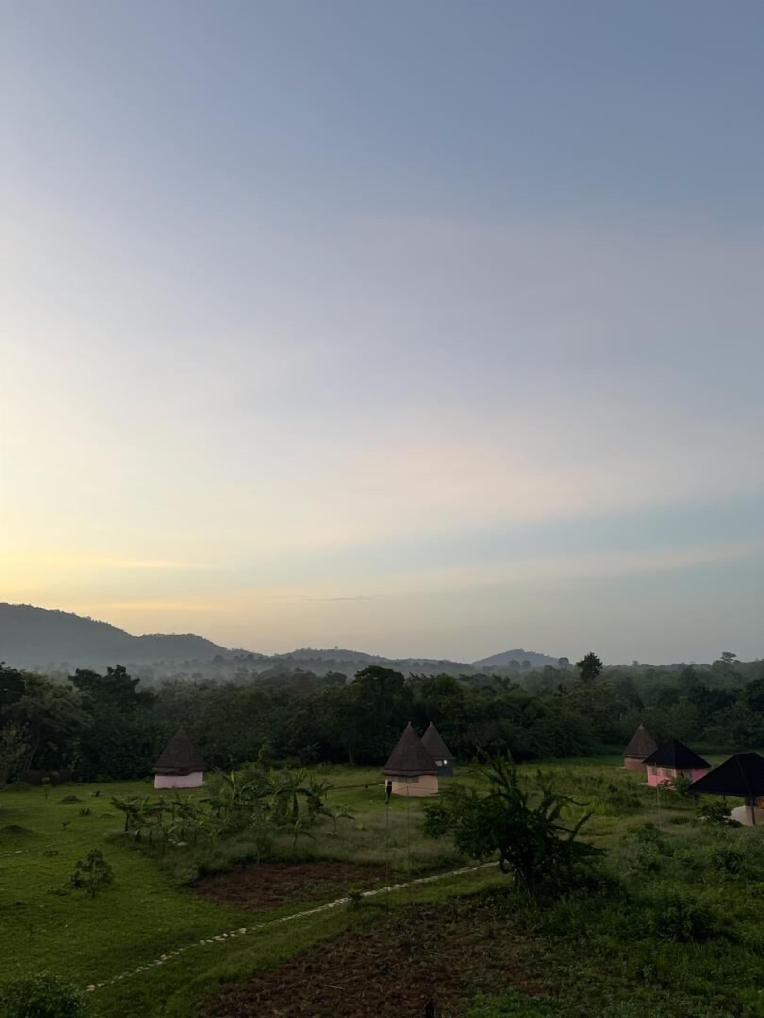 A rural landscape with several small, traditional-style huts with conical roofs, surrounded by lush greenery and hills in the background.