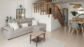 Una sala de estar moderna con un sofá gris, una escalera de madera, una mesa de comedor con sillas y una mesita con una lámpara.