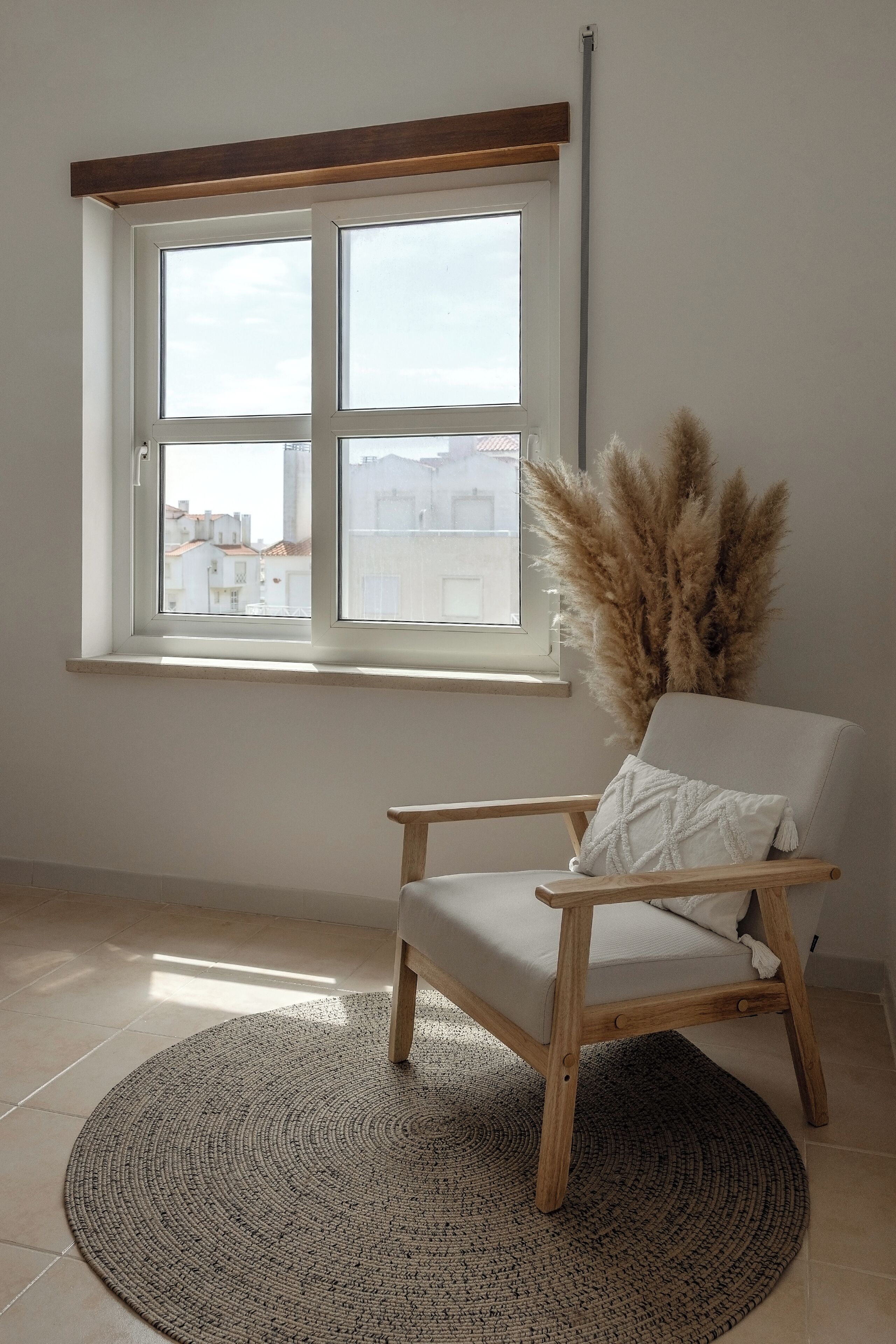 Een kamer met een groot raam, een houten stoel, een geweven vloerkleed en pampasgras decoratie.