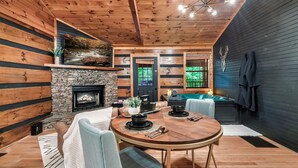 A cozy cabin interior with a stone fireplace, wooden walls, a dining table with chairs, and a view of the outdoors.