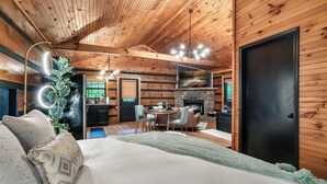 A cozy cabin interior with a wooden ceiling, a dining area, a fireplace, and a bed with a green throw blanket.