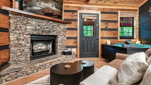 A cozy cabin interior with a stone fireplace, wooden walls, a dark door, and a seating area with a round table and cushions.