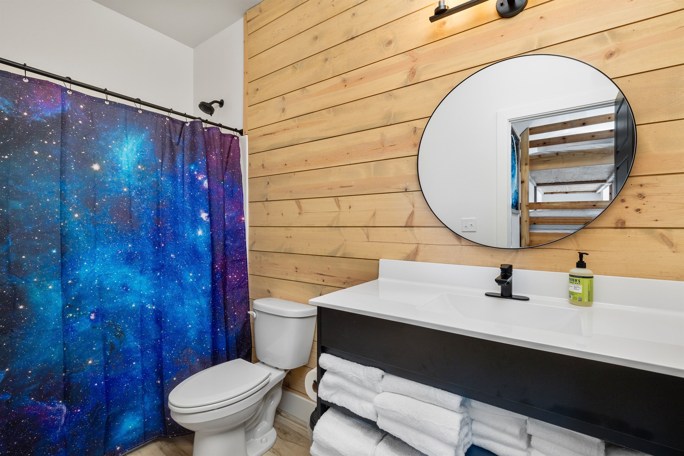 A bathroom with a galaxy-themed shower curtain, a white toilet, a wooden wall, a round mirror, and a white sink with a soap dispenser.