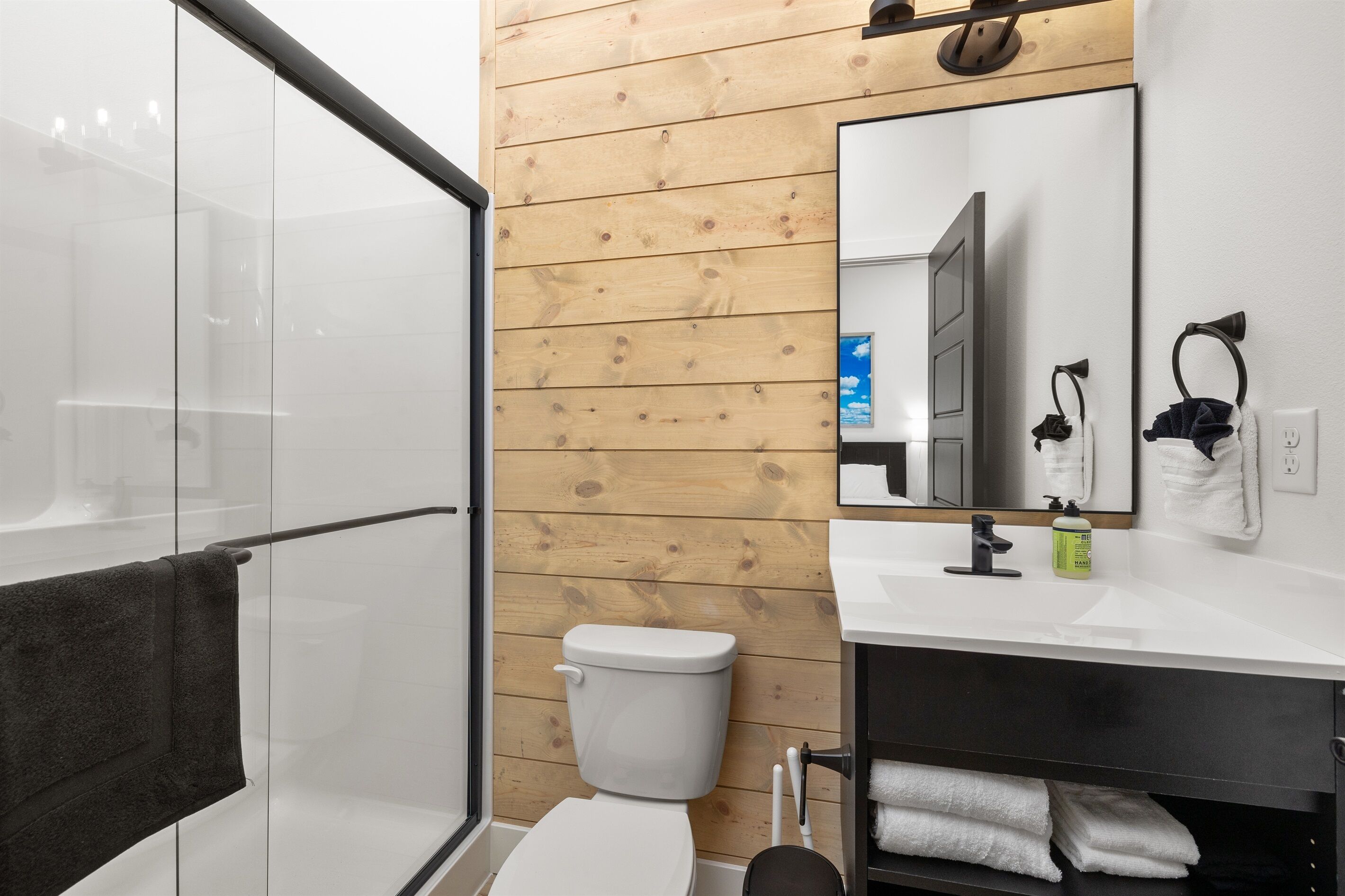 Une salle de bain moderne avec un mur recouvert de panneaux en bois, une douche en verre, des toilettes blanches, un lavabo blanc avec un miroir et un porte-serviettes noir.