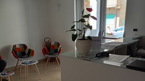 A modern living room with a white desk, two colorful chairs, a potted plant, and a window with a view of the outside. - Hotel Riviera  (Silvi)