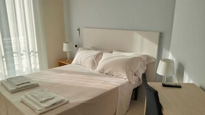 A neatly made bed with a white comforter and pillows, a bedside table with a lamp, and a chair with a remote control. - Hotel Riviera  (Silvi)
