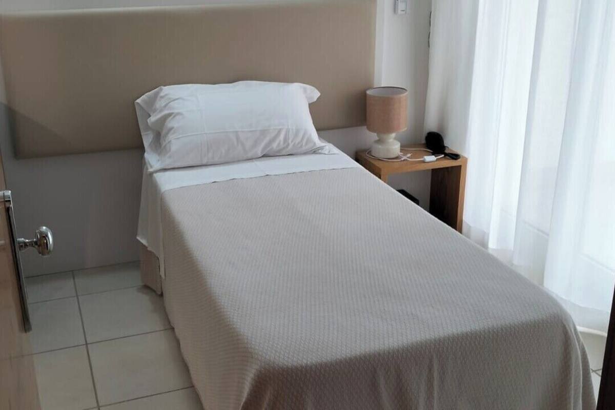 A neatly made bed with a white bedspread and pillows, a bedside table with a lamp, and a door to the left.