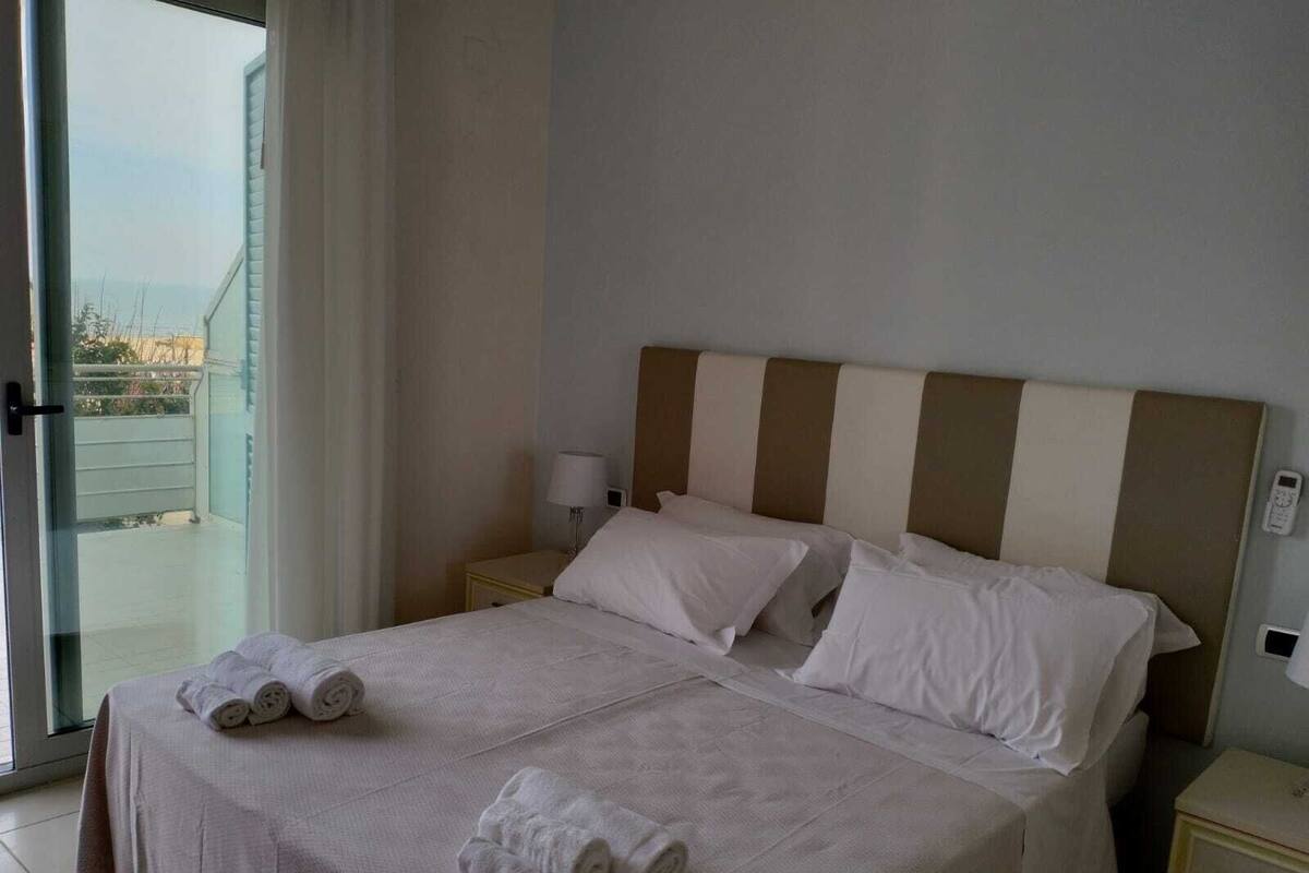 A hotel room with a bed, a nightstand, a lamp, and a view of the outside.