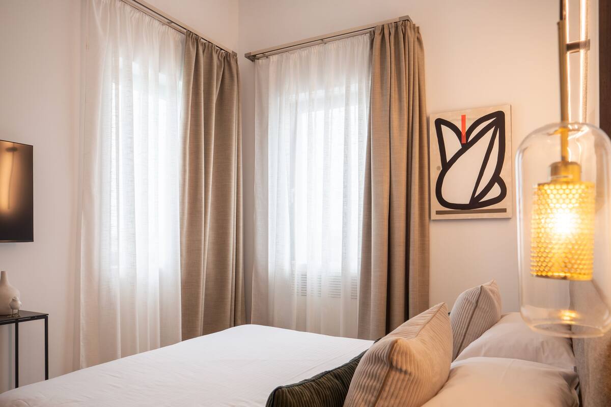 Una habitación de hotel con cama, cortinas, una obra de arte colgada en la pared y una lámpara.