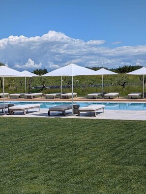 Un área junto a la piscina con sillas de descanso y sombrillas, cielo despejado y césped al frente.
