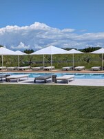 Un área junto a la piscina con sillas de descanso y sombrillas, cielo despejado y césped al frente.