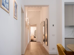 A hallway with a wooden floor, white walls, and framed artwork. There is a door leading to a room with a bed and a mirror. - Tenuta Asmara Luxury Resort (San Miniato)
