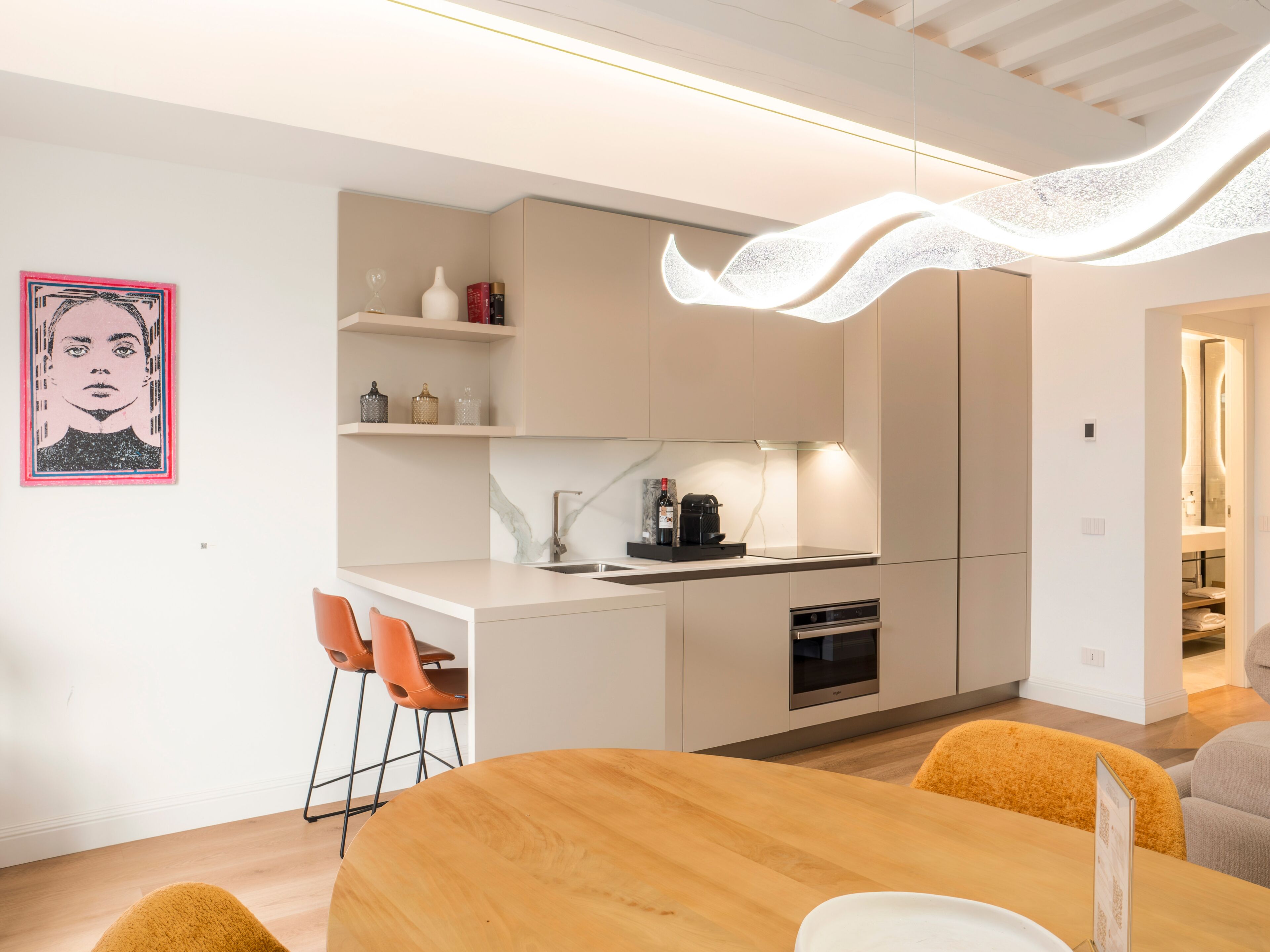 Una cocina moderna con armarios blancos, encimera de mármol y horno empotrado. Hay dos sillas anaranjadas a una mesa de comedor de madera.