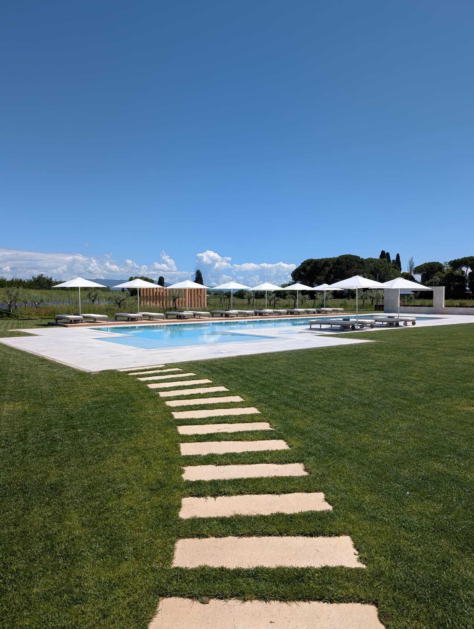 Une piscine sous un ciel dégagé, une zone herbeuse avec des pas japonais, et plusieurs parasols blancs.