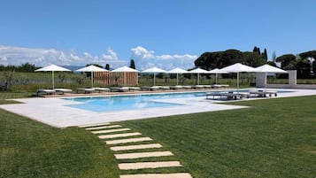 Une piscine sous un ciel dégagé, une zone herbeuse avec des pas japonais, et plusieurs parasols blancs.