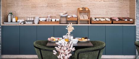 A breakfast setup with a wooden counter, a seating area with green upholstered chairs, and a vase with dried flowers.