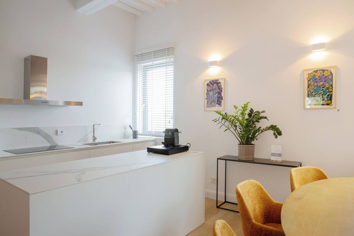 Una cocina moderna con estufa integrada, un lavamanos y una cafetera. Hay una pequeña mesa con una planta y un cuadro en la pared.