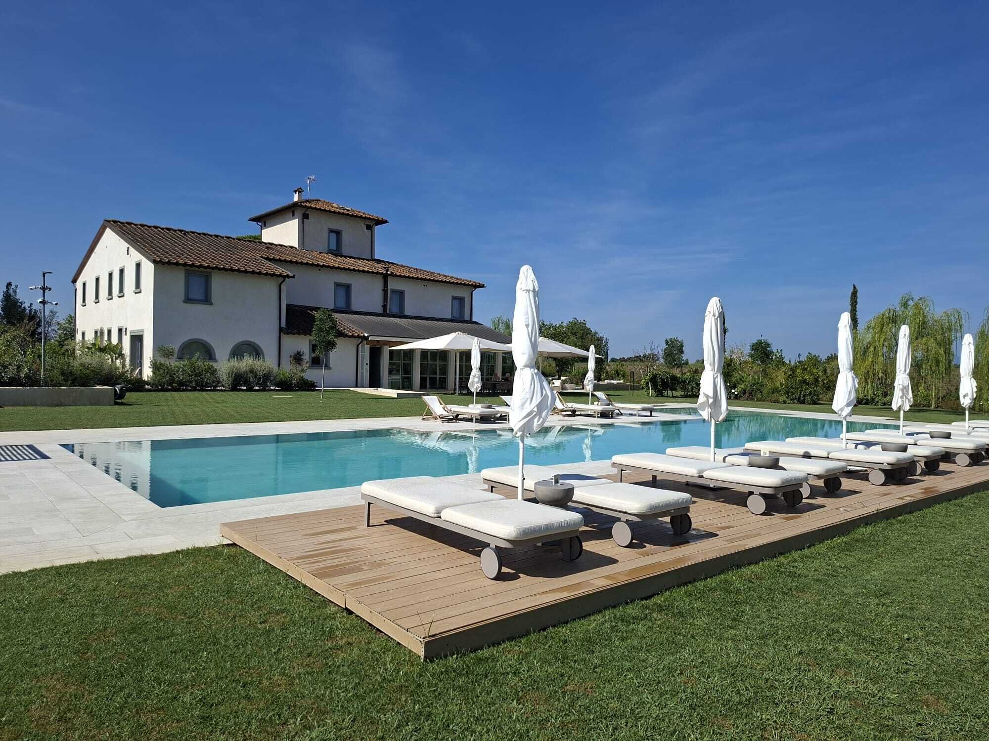 Area piscina con lettini e ombrelloni, cielo azzurro intenso e una grande casa sullo sfondo.