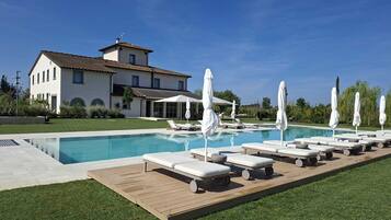 Area piscina con lettini e ombrelloni, cielo azzurro intenso e una grande casa sullo sfondo.