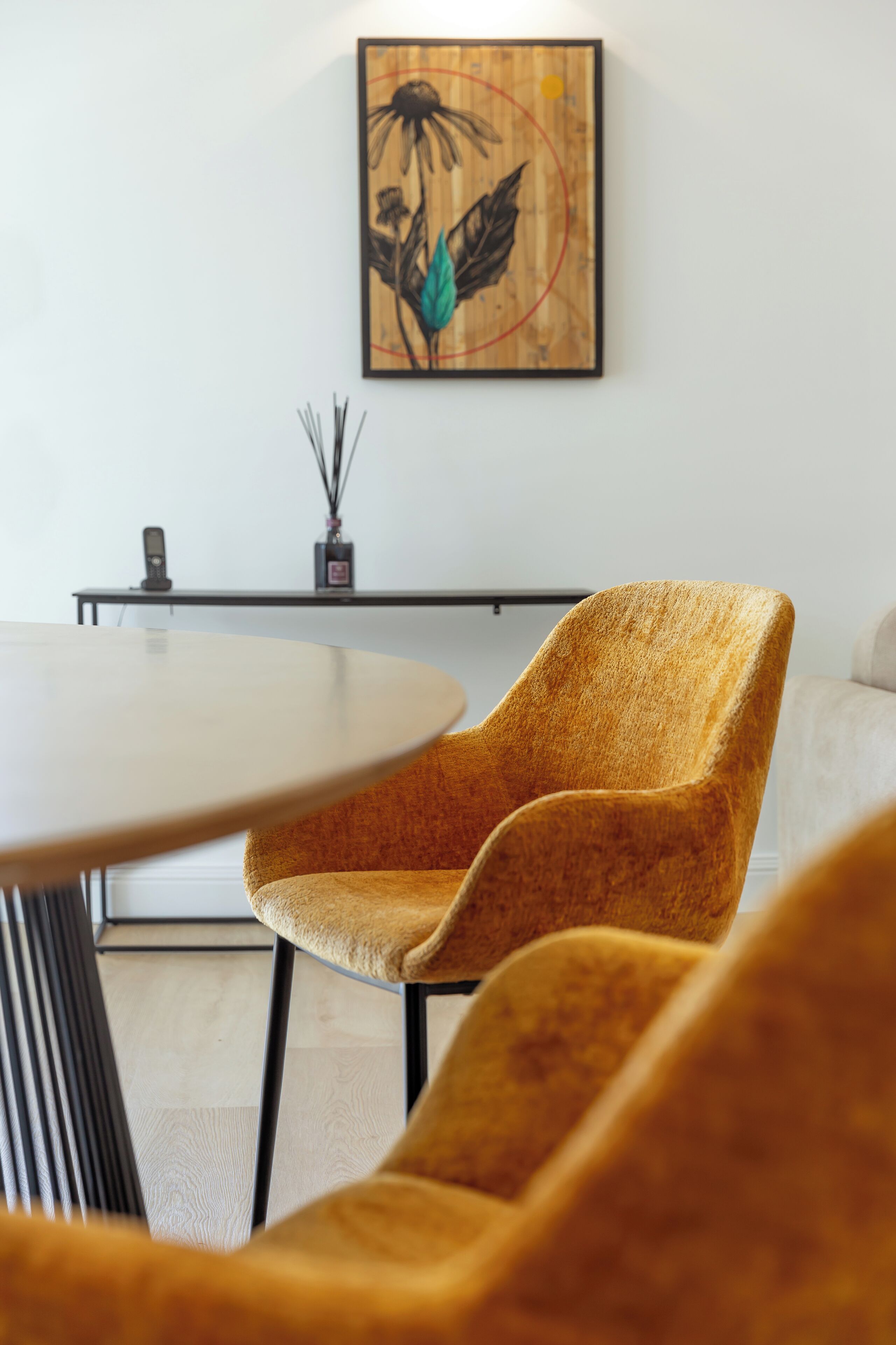 A modern dining area with a round table, two upholstered chairs, a wall-mounted artwork, and a small table with a vase and candle.