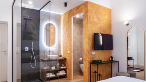 A modern bathroom with a glass shower enclosure, a large mirror, a toilet, and a TV mounted on the wall. - Tenuta Asmara Luxury Resort (San Miniato)