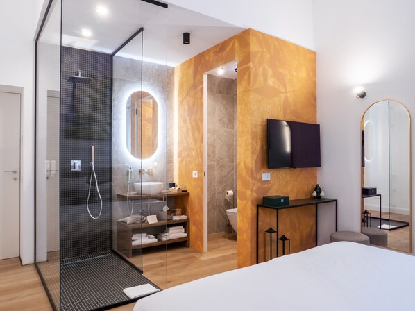 A modern bathroom with a glass shower enclosure, a large mirror, a toilet, and a TV mounted on the wall. - Tenuta Asmara Luxury Resort (San Miniato)