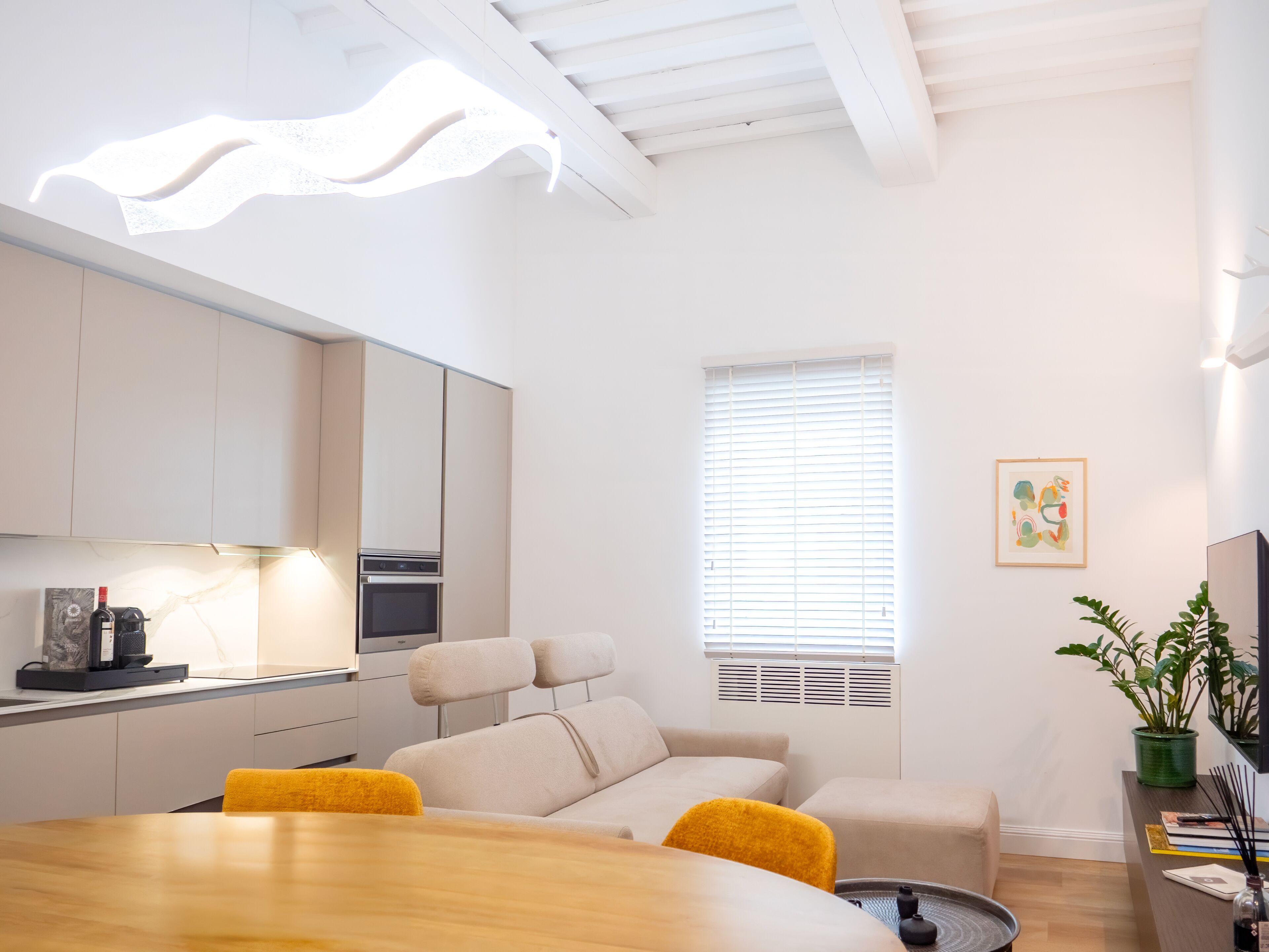 A modern living room with a wooden dining table, a sofa, and a window with blinds.