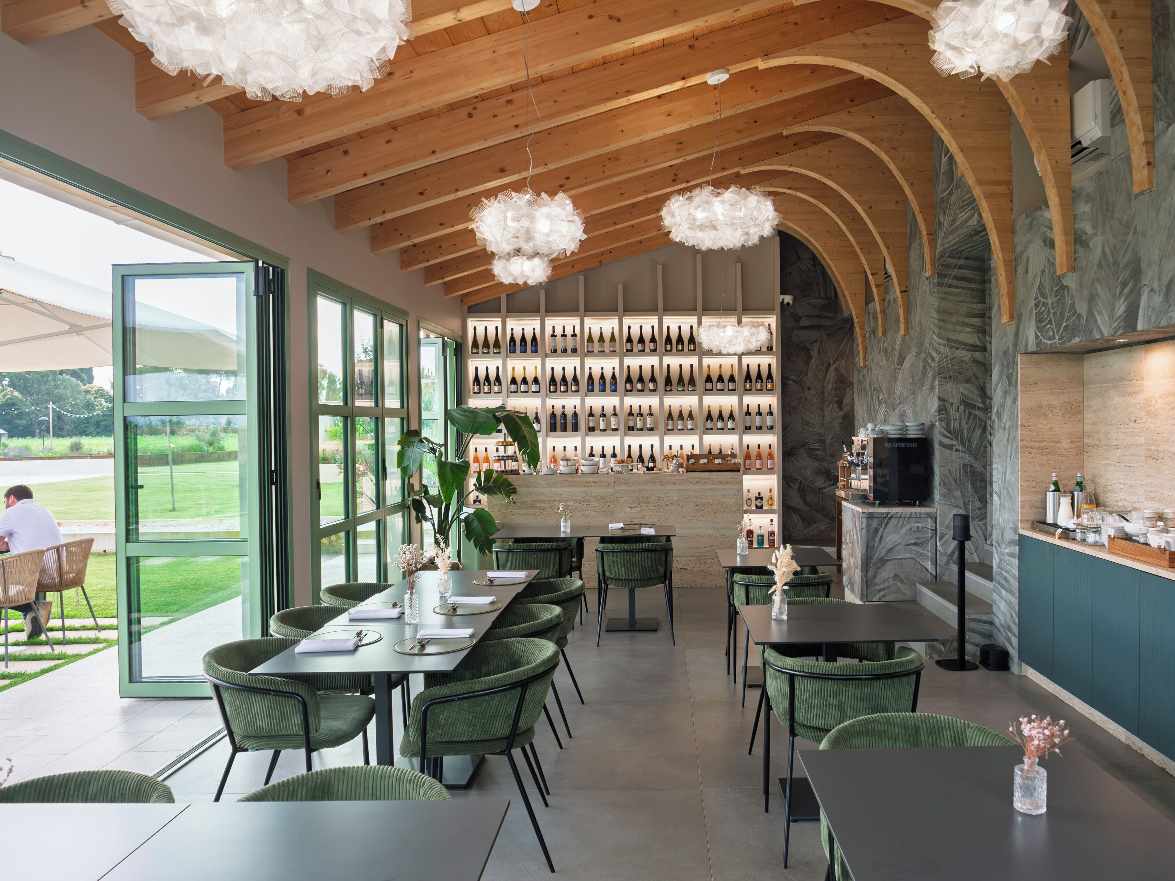 Interior de un restaurante con una larga mesa de comedor, sillas verdes, una pared de vinos y una zona de bar.