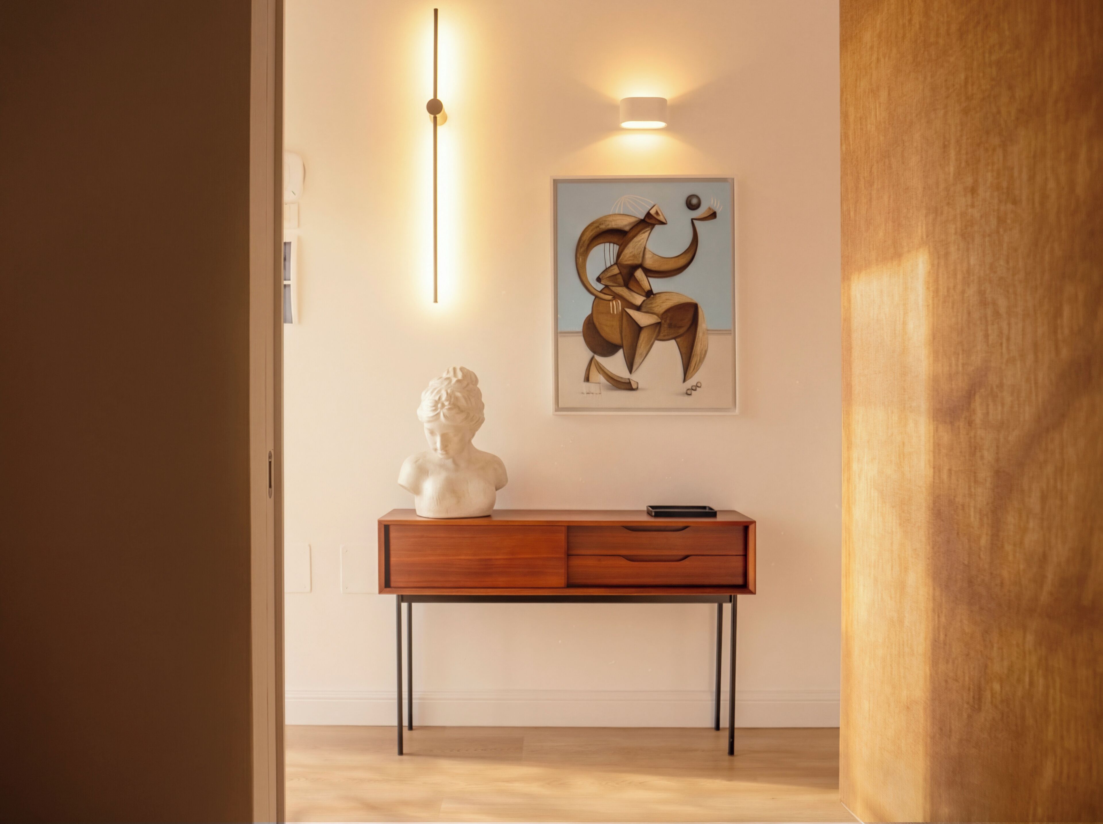 Una habitación con una consola de madera, un busto, un cuadro abstracto enmarcado y un aplique de pared.