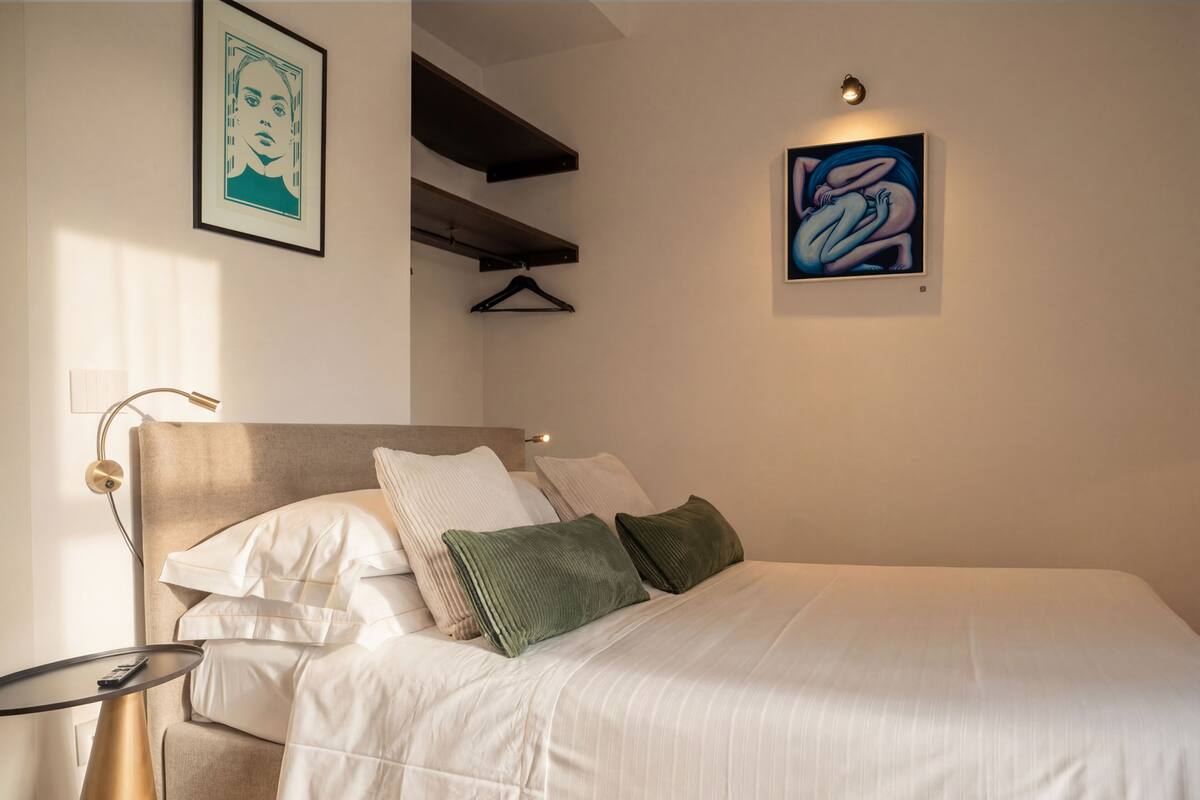 Un dormitorio ordenado con cama, almohadas, mesita de noche, lámpara y obras de arte en las paredes.