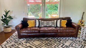 A brown leather sofa with patterned cushions, a large patterned rug, a potted plant, and a guitar in a room with a view of greenery through a glass door.