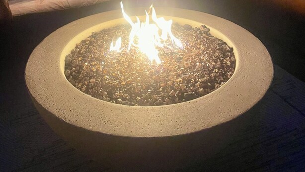 A circular fire pit with a fire burning in the center, surrounded by a concrete ring. There is a sofa with cushions and a hanging macrame light fixture in the background.
