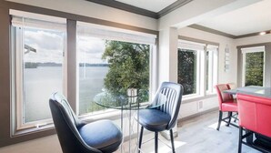 Ett modernt matområde med ett glasbord och stolar, stora fönster med utsikt över en sjö och en röd bardisk.