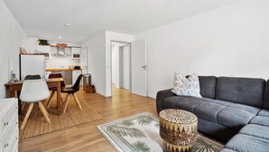 Un salon moderne avec un canapé gris, un coin repas avec une table et des chaises en bois, et une cuisine avec des meubles blancs et un four à micro-ondes.