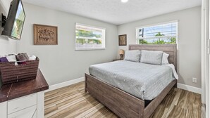 Une chambre à coucher avec un lit en bois, un téléviseur à écran plat fixé au mur et deux fenêtres équipées de stores.