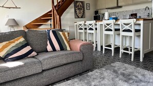 Ett mysigt vardagsrum med en grå soffa, randiga kuddar, en mjuk matta och en trätrappa som leder upp till ett kök med vita stolar och ett kaklat stänkskydd.