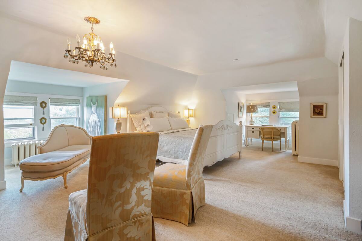 A spacious bedroom with a large bed, a sitting area with a chair, a desk, and a chandelier.