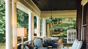 Un portico coperto con colonne bianche, mobili da esterno e vista su un giardino e un edificio sullo sfondo.