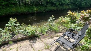 Una sedia vicino a un fiume, con vegetazione lussureggiante e una struttura in legno nelle vicinanze.